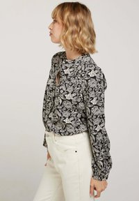 Blouse noire à fleurs avec des manches longues et un décolleté en forme de trou de serrure. Tissu doux présentant des motifs botaniques blancs. Assortie à un pantalon de couleur claire.
