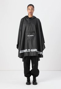 Svart vattentät poncho med huva, med en framficka, vit text "KARL LAGERFELD" på nederkanten, och en lös passform.