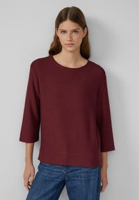 Top lungo bordeaux a maniche lunghe con texture a coste e scollatura rotonda, abbinato a jeans blu. Il design è comodo e casual.