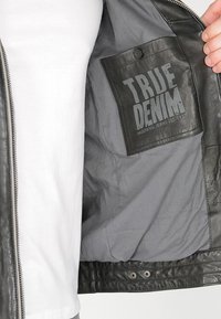 Svart skinnjacka med grå foder, med en ficka märkt "TRUE DENIM" i fet, kontrasterande bokstäver. Dragkedjestängning och knappdetalj.