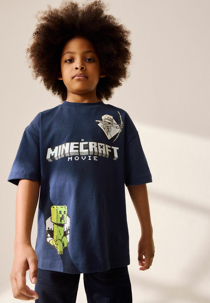 Kind met krullend haar staat en draagt een marineblauw Minecraft film T-shirt met een creeper en kruisboog ontwerp.