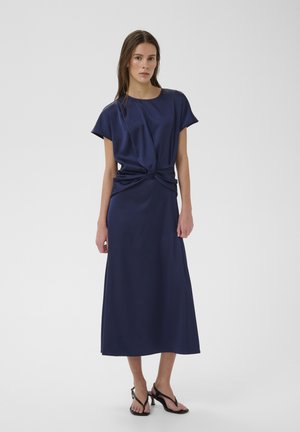 Naine sinimustas midi-kleidis, millel on sõlmevöökoht, koos mustade kontsakingadega, ühevärvilise tausta ees.