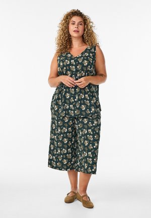 Femme aux cheveux bouclés portant un top sans manches fleuri vert foncé et un pantalon large court assorti, debout, les mains jointes.