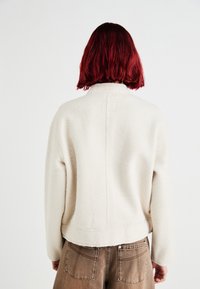 Beige, struktureret sweater med høj hals og sænkede skuldre. Bagpå har den en midtersøm og sidder over brune, højhældte bukser.