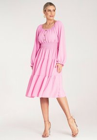Różowa sukienka z długim rękawem, marszczonym pasem, warstwową spódnicą i detalami w postaci guzików. Materiał ma gładką fakturę, dopełniony cielistymi szpilkami.