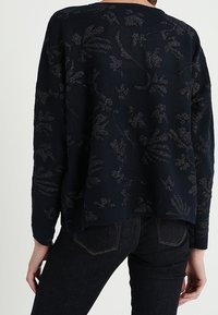 Mörkblått stickat tröja med ett blommönster i en ljusare nyans, med rund halsringning och avslappnad passform. Bärs med mörka denimjeans.