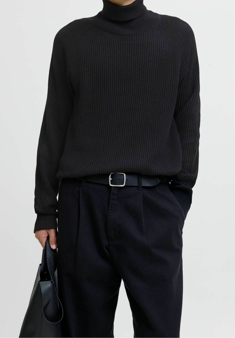 Pull à col roulé côtelé noir à manches longues, associé à un pantalon sombre et une ceinture noire. Un sac fourre-tout noir est tenu dans la main droite.