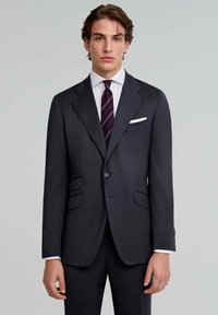 Chaqueta de traje gris oscuro, con solapas de muesca, dos bolsillos frontales, cierre de un solo botón, combinada con una camisa blanca y una corbata a rayas.