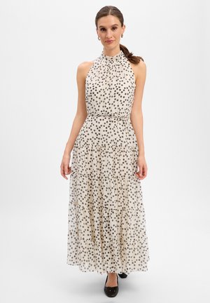 Frau trägt ein ärmelloses, hochgeschlossenes, knöchellanges weißes Kleid mit schwarzen Polka-Dots und einem dünnen Taillengürtel, kombiniert mit schwarzen Ballerinas.