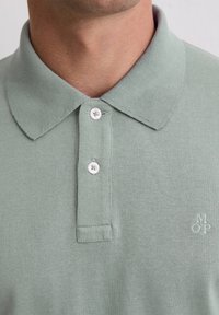 Cămașă polo verde deschis, cu o textură moale, cu un guler clasic și un închizător cu trei butoane, având un mic logo brodat pe piept.