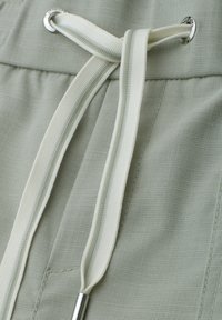 Gros plan sur un pantalon en tissu vert pâle avec un cordon blanc passé dans des œillets métalliques à la taille.