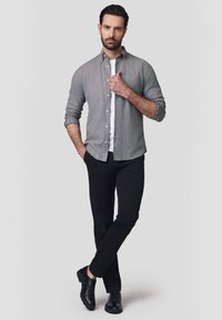 Szara koszula zapinana na guziki na białym t-shircie, w połączeniu z czarnymi spodniami i czarnymi butami. Model stoi pewnie na tle w neutralnych kolorach.