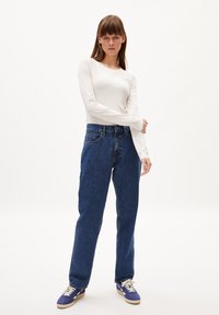 Weißes Langarmhemd, blaue High-Waist-Jeans und lila Turnschuhe. Die Jeans haben eine gerade Passform und ein Fünf-Taschen-Design.