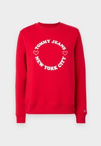 Sudadera roja de manga larga con texto circular blanco "TOMMY JEANS NEW YORK CITY" y dos gráficos de corazones rosa en la parte frontal.
