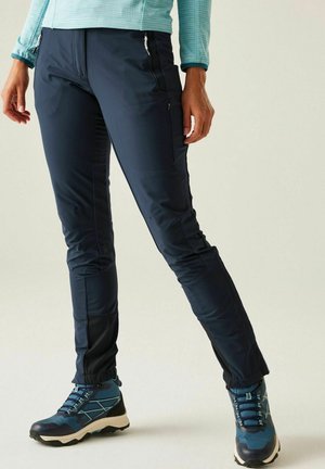 Person trägt marineblaue Outdoor-Hose mit Reißverschlusstaschen und blaue Wanderschuhe, steht mit überkreuzten Beinen vor einfachem Hintergrund.