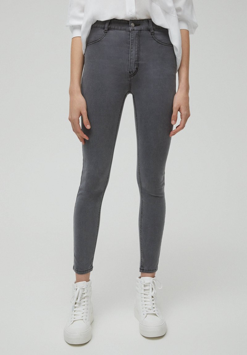Jeans ajustados en denim negro desgastado, con cintura alta, dos bolsillos frontales y una textura suave. Llevados con zapatillas blancas de caña alta.