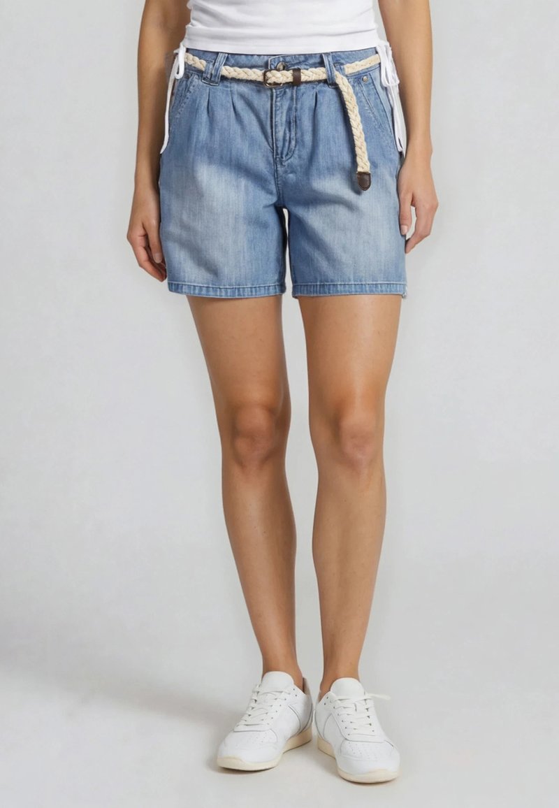 Personne portant un short en denim bleu clair avec une ceinture beige tissée, un haut blanc et des baskets blanches, debout devant un fond uni.