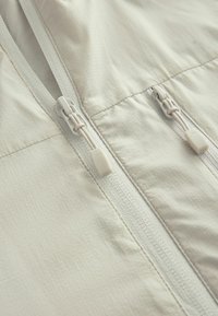 Light beige zippered jacket featuring a textured fabric, dual zippers with pull tabs, and sleek stitching along the seams.