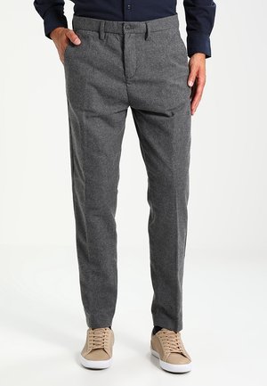 Mand iført skræddersyede grå bukser med forlommer og beige sneakers, stående mod en hvid baggrund.