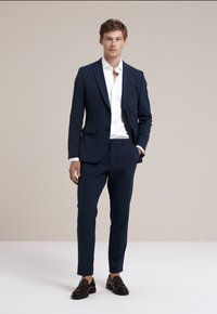 Costume bleu marine avec blazer ajusté et pantalons slim, associé à une chemise blanche et des mocassins en cuir marron. Texture de tissu lisse.