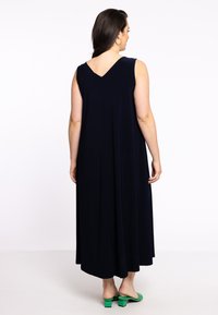 Marineblaue, ärmellose Maxikleid mit V-Ausschnitt-Design, das eine fließende Silhouette zeigt. Das Model trägt grüne, hochhackige Mules.
