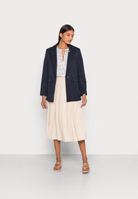Blazer azul marinho com gola notched, emparelhado com uma blusa leve estampada e uma saia midi plissada bege. Sandálias brancas com tiras completam o visual.