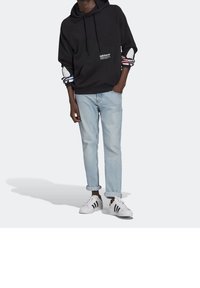 Svart hoodie med vitt logotyp, mönstrade ärmslut i röd och blå, kombinerad med ljusblå upprullade jeans och vita sneakers med svarta ränder.