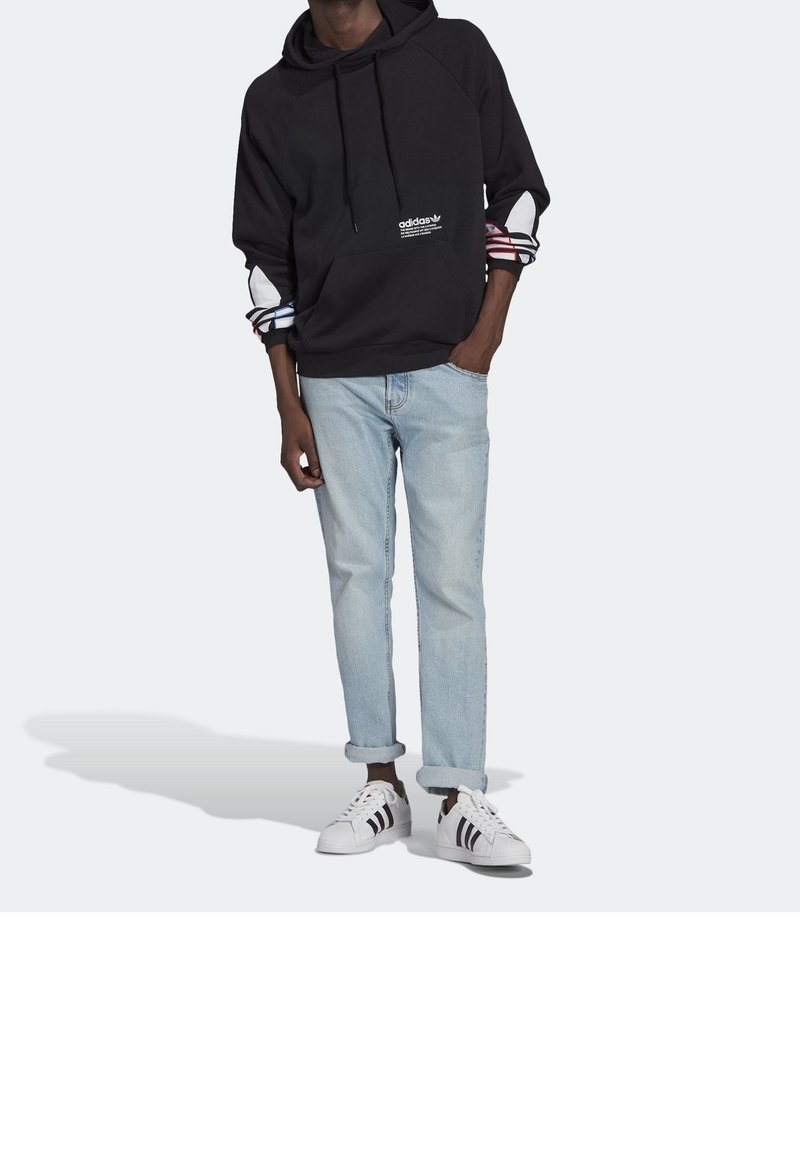 Svart hoodie med vitt logotyp, mönstrade ärmslut i röd och blå, kombinerad med ljusblå upprullade jeans och vita sneakers med svarta ränder.