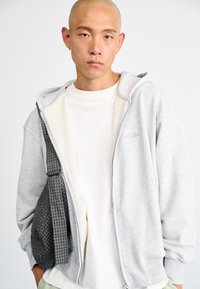 Sivá zipová mikina s kapucňou a bielym interiérom, nosená cez jednoduché biele tričko. Má na pleci karovanú sivú crossbody tašku.