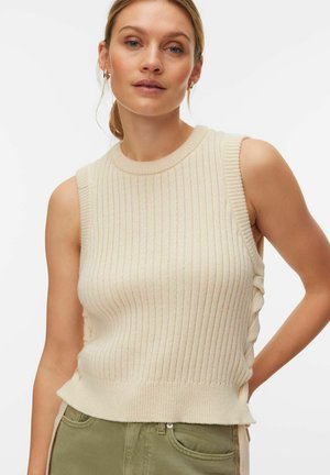 Femme aux cheveux blonds portant un haut sans manches beige en tricot côtelé et un pantalon vert clair, debout sur un fond blanc uni.