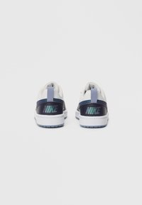 Pereche de adidași Nike albi și bleumarin cu logo iridescent pe călcâi și desfacători de ușă de culoare bleu deschis, văzuți din spate pe un fundal simplu.