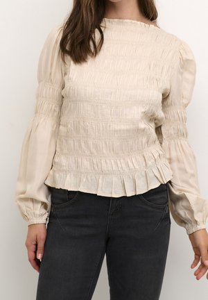 Femme portant un chemisier écru smocké avec de longues manches bouffantes et un jean noir taille haute, debout devant un fond clair.