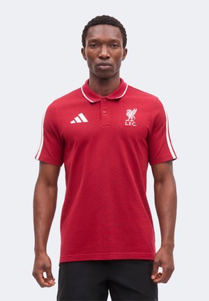 Άνδρας που φορά κόκκινο πόλο μπλούζα της Liverpool FC με λευκό λογότυπο Adidas και ρίγες στα μανίκια, στέκεται μπροστά σε απλό φόντο.