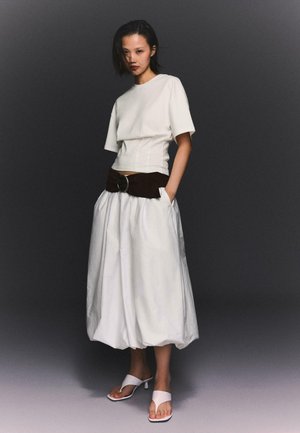 Woman standing with hands in pockets wearing a white short-sleeve top, voluminous white skirt, wide dark belt, and white heeled sandals.