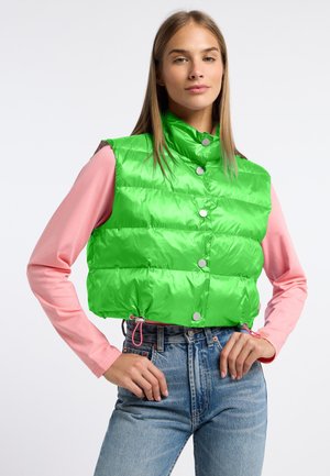 Vrouw met felgroene cropped puffer-vest over een roze shirt met lange mouwen en een high-waisted blauwe jeans, staand met handen in de zij.