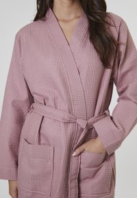 Batín de punto waffle en color rosa, con diseño envolvente, cintura con cinturón, mangas largas y dos bolsillos frontales. Textura suave con un patrón a cuadros.