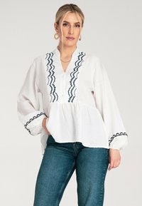 Biała bluzka z niebieskimi haftowanymi zygzakami i okrągłymi wzorami, z dekoltem w serek, długimi rękawami i falbaną w talii. Stylizowana z ciemnymi dżinsami.