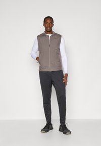Gilet leggero grigio con chiusura a zip, due tasche laterali e colletto, indossato sopra una camicia bianca a maniche lunghe e pantaloni sportivi neri.