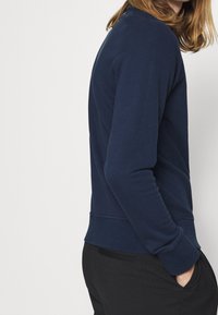 Sudadera de cuello redondo en azul marino, fabricada en una suave mezcla de algodón, con un corte relajado, puños acanalados y dobladillo cosido, combinada con pantalones negros.