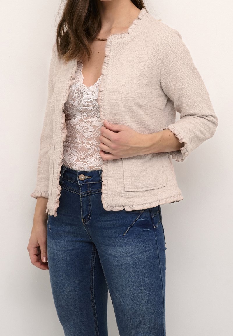 Femme portant un cardigan rose pâle texturé à ouverture devant avec des bords froncés, par-dessus un haut en dentelle blanche et un jean bleu, tenant le devant du cardigan à la main.