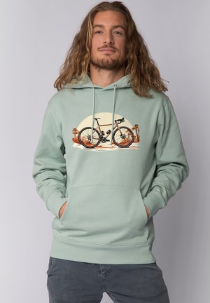 Mann mit langen Haaren trägt einen mintgrünen Hoodie mit einer Grafik eines Kieselradfahrrads und einer Wüstenlandschaft mit Palmen.