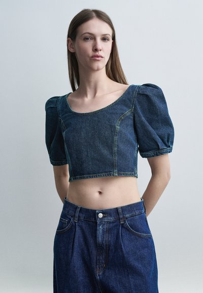 Jeune femme aux cheveux longs portant un haut en denim court à manches ballons et un jean taille haute, debout devant un fond clair uni.