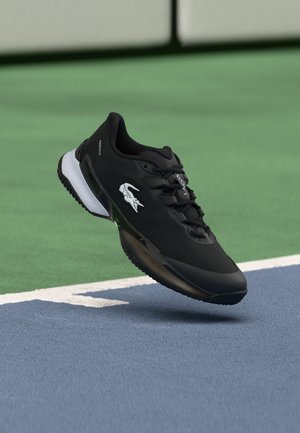 Zapatilla deportiva negra de Lacoste con el logo de cocodrilo blanco, mostrada en el aire sobre una superficie de pista de tenis azul y verde.
