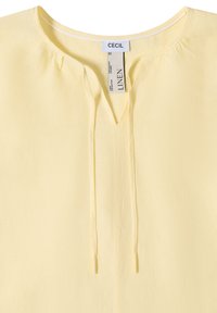 Blouse en lin jaune clair avec un col rond, un détail froncé et des liens sur le devant, étiquetée "CECIL" à l'intérieur du col.