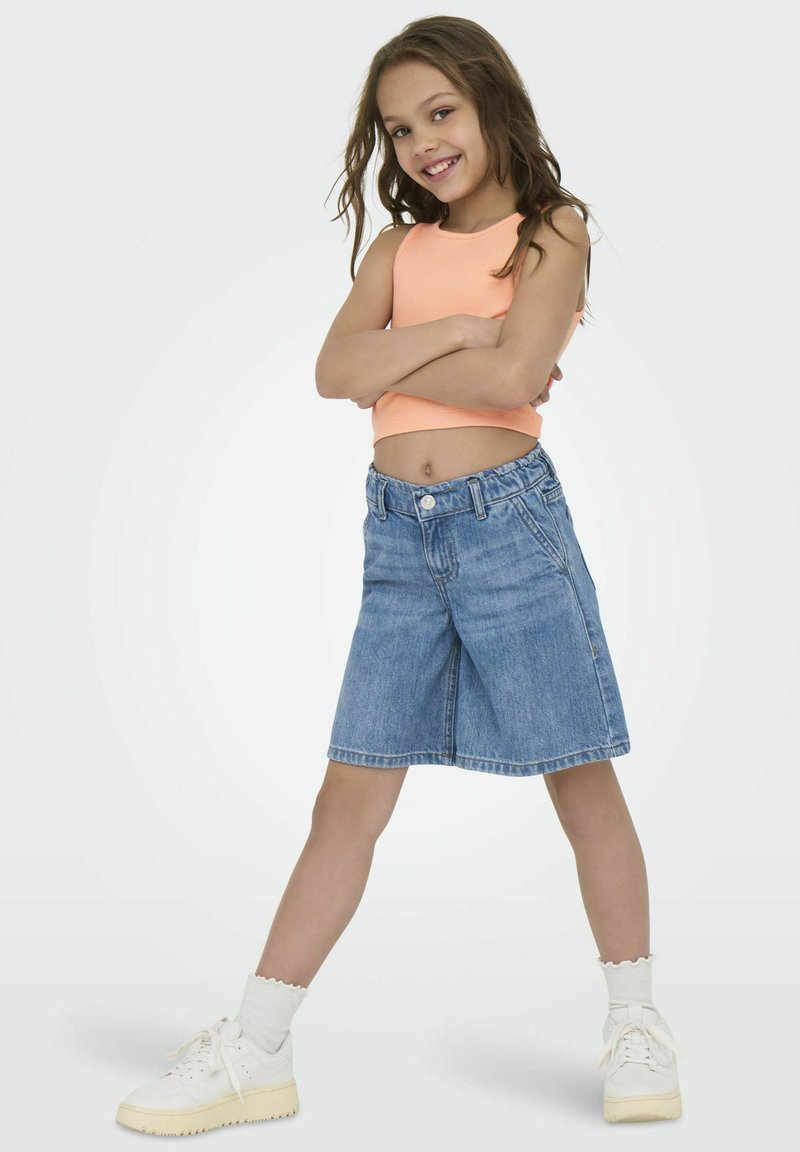 Shorts en denim à taille mi-haute, délavage bleu clair et ourlet roulé ; associés à un crop top orange vif et des baskets blanches.