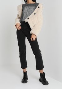 Chaqueta de piel sintética beige con botones negros sobre una camisa gris con estampado, combinada con jeans negros cortos y botines negros con cordones.