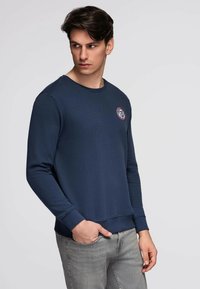 Ung mand iført en marineblå sweatshirt med et lille rundt logo på brystet, lysegrå jeans, stående med den ene hånd i lommen og kiggende mod venstre.