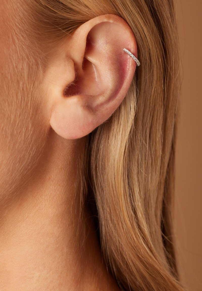 Boucle d'oreille en argent avec de petits gemmes incrustées ; design élégant et arrondi qui s'adapte parfaitement au haut de l'oreille. Cheveux bruns partiellement visibles.