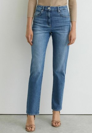 Femme portant un jean droit bleu clair, un haut beige à manches longues côtelé et des sandales beiges à bout ouvert, debout sur un sol gris.