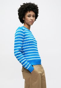 Μπλε ριμπ πλεκτή μπλούζα με έντονες οριζόντιες ρίγες σε ανοιχτό μπλε και λευκό, με στρογγυλή λαιμόκοψη και μακριά μανίκια, σε συνδυασμό με μπεζ παντελόνι.